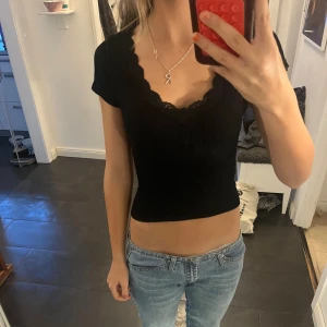 Svart croppad ribbad topp med spets - Säljer en svart croppad topp med ribbad struktur och v-ringad hals med spetskant. Toppen har korta ärmar och sitter tight mot kroppen. Perfekt för dig som gillar en enkel men ändå snygg look med lite extra detalj vid halsen.🌸