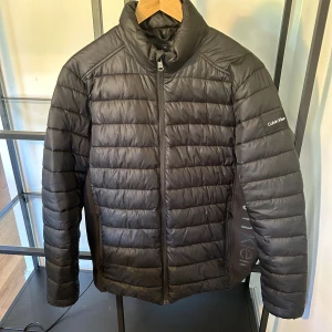 Svart pufferjacka från Calvin Klein M - Svart pufferjacka från Calvin Klein i storlek M. Jackan är quiltad med horisontella sömmar, har hög krage och dragkedja framtill. Tillverkad i 100% återvunnen polyester och har diskret logga på ärmen. Perfekt för kyliga dagar. 