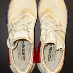Adidas NMD vita sneakers med färggdetalj - Säljer ett par Adidas NMD sneakers i vitt med klassiska tre ränder på sidan. Skorna har en chunky vit sula med röda och blå detaljer på sidorna, samt snörning och rund tå. Ovandelen är i syntetmaterial och har en modern, sportig look.