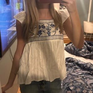 Vit blus med blå broderi - Supersöt vit blus med blått broderat blommönster upptill och små prickar i tyget. Blusen har korta volangärmar och en luftig, flowy passform. Perfekt för varma dagar och ger en romantisk vibe till outfiten.