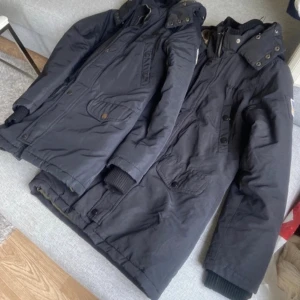 Svart parkasjacka från Svea.tvillingar.S - Säljer en svart parkasjacka från Svea med hög krage, avtagbar huva och flera fickor med både knappar och dragkedja. Jackan har långa ärmar med ribbade muddar och snygga metallknappar. Perfekt för kalla dagar och har en stilren look.har 2 st nästan likadana 750 kr /st .mycket bra skick hämtas i norsborg botkyrka.