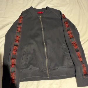 Hugo Boss Hoodie - Snygg svart bomberjacka från Hugo med röda logoband längs ärmarna. Jackan har dragkedja framtill, ribbade muddar och två fickor. Materialet är mjukt och skönt, perfekt för en streetwear-look.