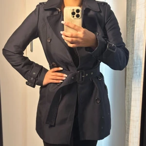 Mörkblå trenchcoat med bälte - Snygg mörkblå trenchcoat med klassisk dubbelknäppning, bälte i midjan och spännen vid ärmsluten. Jackan har bred krage och knappar framtill, perfekt för dig som vill ha en stilren och tidlös look.