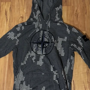 Stone Island grå hoodie med camomönster - Grym grå hoodie från Stone Island med vitt camoinspirerat rutmönster och stor broderad kompasslogga på bröstet. Klassisk huva med snörning och ikonisk patch på ärmen. Mjuk insida i bomullsmix, perfekt för chill dagar.