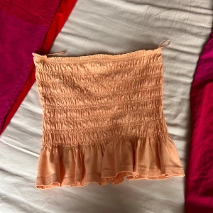 Persikofärgad tubtopp från Gina Tricot - Säljer en söt persikofärgad tubtopp från Gina Tricot i storlek S. Toppen är smockad med volangkant nedtill och har bandeau-modell. Perfekt för varma dagar och har en mjuk, stretchig känsla. Supersnygg att styla med jeans eller kjol!