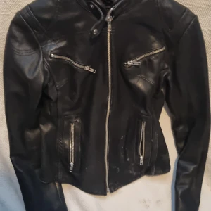 Svart skinnjacka Camie Hoxton London - Snygg svart skinnjacka från Camie Hoxton London med coola detaljer som flera dragkedjor framtill och på ärmarna. Jackan har figurnära passform och lång ärm, perfekt för dig som gillar en edgy stil. Klassisk bikerlook med hög krage och silverfärgade metalldetaljer.