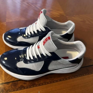 Prada sneakers i blått och vitt - Snygga Prada sneakers med vit mesh och blanka mörkblå detaljer. Skorna har vita snören, rund tå och en vit sula. Röd Prada-logga på plösen ger en lyxig touch. Perfekta för dig som vill ha en sportig men exklusiv look.