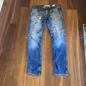 Blå slitna jeans från Replay - Säljer ett par blå jeans från Replay med coola slitningar och trasiga detaljer på framsidan. Jeansen har klassisk femficksdesign, raka ben och tydliga tvättade partier som ger en grymt avslappnad vibe. Perfekt för dig som gillar streetstyle.