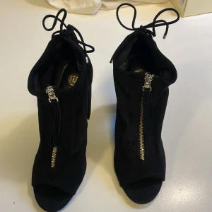 Svarta stövletter från River Island - Svarta stövletter i mocka-look från River Island med chunky hög klack, öppen tå och dragkedja framtill. Snygga snördetaljer bak med metalltips. Perfekta för dig som vill sticka ut med en edgy vibe.