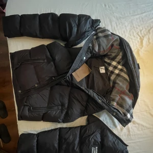 Svart pufferjacka från Burberry/bb storlek S / P - Svart pufferjacka från BB med klassiskt rutigt foder i grått, vitt och rött. Jackan har dragkedja, stora fickor med dragkedja och tydlig Burberry-logga på ärmen. Perfekt för kalla dagar och riktigt snygg streetstil.  Jackan är helt oanvänd och säljs för den inte passar.
