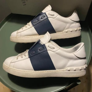 Valentino Garavani Open sneakers vit/blå - Snygga Valentino Garavani Open sneakers i vitt skinn med bred marinblå läderdetalj över sidan. Klassisk rund tå, platt sula och snörning framtill. Ikoniska nitar längs hälen ger extra edge. Perfekt för dig som gillar lyxiga och stilrena sneakers. Snörerna är rengjorda.