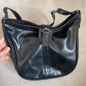 Svart axelväska - Snygg svart axelväska med justerbart axelband och silverfärgat spänne framtill. Väskan har dragkedja på baksidan och flera fack inuti, Enkel och stilren design som passar till det mesta. Köpt second hand.