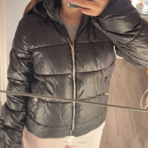 Svart pufferjacka från ONLY med huva - Säljer en svart pufferjacka från ONLY med huva och dragkedja framtill. Jackan har quiltad design, två fickor med tryckknappar och en snygg logga på ärmen. Perfekt för kalla dagar och riktigt skön att bära.