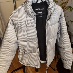 Randig pufferjacka från FB Sister XS - Snygg pufferjacka från FB Sister i storlek XS. Jackan är vit med svarta tunna ränder, har dragkedja framtill och två fickor. Perfekt för dig som vill ha en cool och trendig look under kyliga dagar.