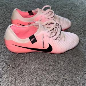 Nike rosa fotbollsskor med snörning - Säljer ett par snygga Nike fotbollsskor i ljusrosa och vitt med svart swoosh på sidan. Skorna har snörning, rund tå och platt sula med dobbar för konstgräsplan. Ovandelen är i syntetmaterial och insidan är rosa. Perfekt för dig som har en bred fot.