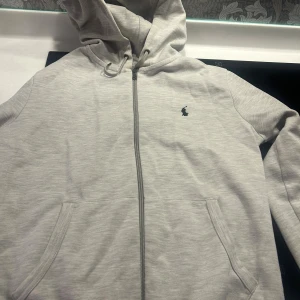 Grå hoodie från Polo Ralph Lauren XS - Snygg ljusgrå hoodie från Polo Ralph Lauren med dragkedja, huva med snörning och klassisk broderad logga på bröstet. Mjuk bomullsblandning och två fickor framtill. Perfekt för chill dagar eller när du vill ha en clean look.