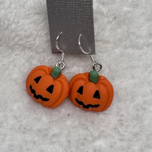 Halloween örhängen med pumpor - Unika örhängen formade som orange pumpor med gröna stjälkar och svarta ansikten, perfekta för Halloween. De har en lekfull design och hängs i silverfärgade krokar. Perfekt accessoar för dig som vill sticka ut under hösten!