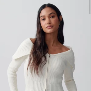 Vit ribbad offshoulder kofta med dragkedja - Säljer en vit ribbad kofta med offshoulder-design och lång ärm. Tröjan har en bred krage som vilar över axlarna och en snygg dragkedja framtill. Modellen är croppad och sitter snyggt på kroppen, perfekt för dig som gillar trendiga detaljer. Typ helt ny! Endast använt ca 2-3 gånger. Ny pris: 400kr.