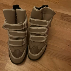 Isabel marant sneakers - Säljer ett par beige höga sneakers från Isabel marant. med flera kardborreband och coola vita detaljer. Skorna har en svart sula och en lila detalj på sidan.