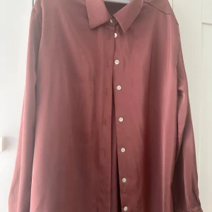 Vinröd skjorta i satin med knappar - Snygg vinröd skjorta i satinliknande material med klassisk krage och vita knappar framtill. Skjortan har lång ärm och en loose passform som ger en avslappnad men stilren look.