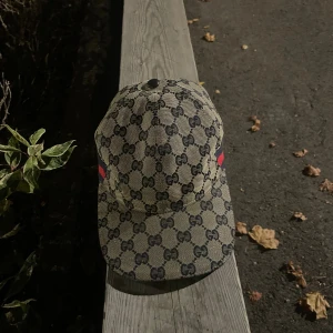 Gucci keps med GG-mönster och röd-blå detalj - Säljer en snygg Gucci keps i beige med klassiskt GG-monogram över hela kepsen. Kepsen har justerbart spänne bak i svart skinn och röd-blå rand på sidan. Tillverkad i Italien och i bomullsmaterial. Perfekt för dig som vill sticka ut med en lyxig accessoar.