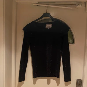 Svart glittrig topp med volang från Skill - Svart långärmad blus från Skill med glittrigt tyg och snygga volangdetaljer vid axlarna. Blusen är helt ny och oanvänd. Storlek 170