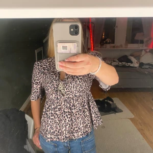 Leopardmönstrad blus med knyt - Säljer en luftig blus från Vero Moda med leopardmönster i beige, svart och rosa toner. Blusen har korta ärmar, volang nedtill och två snygga knytningar framtill som ger en cool cut-out detalj. Perfekt för dig som gillar att sticka ut med djurmönster!