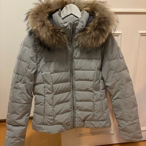 Ljusgrå pufferjacka med fuskpäls från Bomboogie - Säljer en ljusgrå pufferjacka från Bomboogie med huva och fluffig fuskpäls runt kragen. Jackan har quiltad design och dragkedja framtill. Perfekt för kalla dagar och riktigt snygg till vintern. Storlek 3 motsvarar S. 
