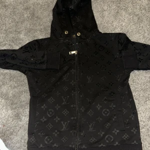 Svart Louis Vuitton hoodie med monogram - Säljer en svart hoodie från Louis Vuitton med ikoniskt monogrammönster över hela tröjan. Tröjan har dragkedja framtill, huva med metallöglor och ribbade muddar. Perfekt för dig som vill sticka ut med en lyxig streetstyle. Sitter litet