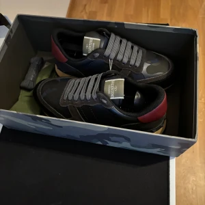 Valentino Garavani Rockrunner sneakers - Snygga Valentino Garavani Rockrunner sneakers i svart och mörkgrått med kamouflagemönster och röda detaljer på hälen. Skorna har grå snörning, platt sula och är tillverkade i en mix av skinn och mocka. Perfekta för dig som vill sticka ut med exklusiva detaljer.