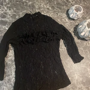 Svart spetsblus med volang från Sisters Point - Supersnygg svart spetsblus från Sisters Point med hög krage och långa ärmar. Blusen har en volangdetalj över bröstet och är helt i spets med transparent känsla. Perfekt för dig som gillar detaljer och vill sticka ut lite extra.