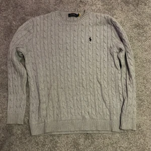 Grå kabelstickad tröja Polo Ralph Lauren - Kabelstickad grå tröja från Polo Ralph Lauren med klassisk rund halsringning och broderad logga på bröstet. Långärmad modell i mjuk bomull, perfekt för lager-på-lager, storlek M.