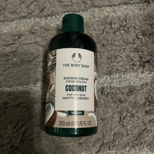 The body shop - Bilden visar en flaska The Body Shop Coconut Shower Cream, 250 ml, för torr hud. Produkten är vegansk, har en nötig och krämig doft och är avsedd för skonsam rengöring. Flaskan ser ut att vara i gott skick utan synliga defekter.