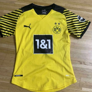 Borussia Dortmund gul matchtröja Puma - Gul Borussia Dortmund fotbollströja från Puma med svarta detaljer och ränder på ärmarna. Kortärmad modell med klubbmärke och sponsortryck på bröstet, samt DORTMUND tryckt på ryggen. Tillverkad i lätt och ventilerande polyester.