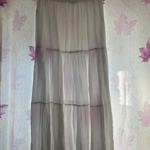 Ljusgrå långkjol i luftig bomull - Säljer en ljusgrå långkjol med volanger och resår i midjan. Kjolen är tillverkad i ett tunt och luftigt bomullstyg som ger en skön och flowy känsla. Perfekt för dig som gillar en avslappnad och stilren look.