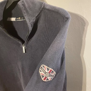 Mörkblå stickad half zip från Henri Lloyd - Mörkblå stickad tröja från Henri Lloyd med half zip och hög krage. Tröjan har ett broderat märke på ena ärmen med brittisk flagga och text. Perfekt för kyligare dagar och passar till många olika stilar.