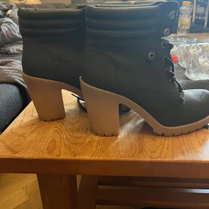 Svarta boots med klack från VOX - Säljer ett par svarta boots från VOX i storlek 38. Skorna har grov klack, snörning framtill och en cool, chunky sula i beige. Perfekta för dig som vill ha en edgy stil med lite höjd. Materialet är syntet och de har en rund tå.