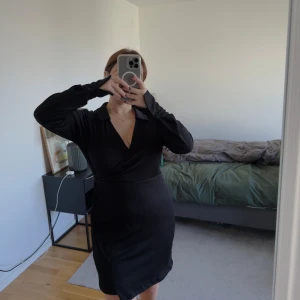 Svart omlottklänning från Bik Bok  - Svart kort omlottklänning från Bik Bok med v-ringning, krage och långa ärmar med breda manschetter. Klänningen är tillverkad i ett lätt strukturerat tyg av polyester och elastan, vilket ger en bekväm passform och snygg siluett.