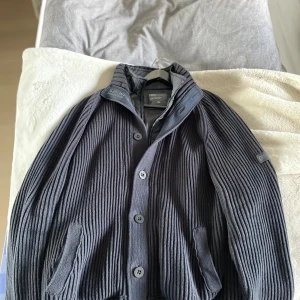 Mörkgrå ribbad cardigan jacka Next  - Riktigt snygg mörkgrå cardigan jacka från Next i storlek XL. Jackan är ribbstickad med ståkrage, knäppning framtill och två fickor. Insidan har quiltad detalj vid kragen och en cool patch på ärmen. Perfekt för dig som gillar stilren och modern look.