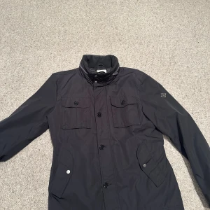 Svart parkajacka från J.Lindeberg - Svart parkajacka från J.Lindeberg med hög krage, fyra framfickor med knappar samt snygga detaljer på ärmsluten. Jackan har en stilren look och är tillverkad i ett slitstarkt syntetmaterial. Perfekt för dig som gillar klassisk och clean stil. Nypris 2300