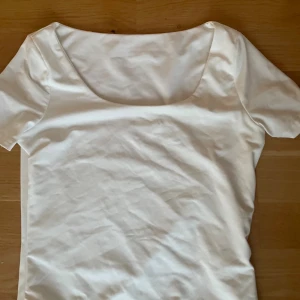 Vit kortärmad topp med rundad urringning - Säljer en stilren vit topp med korta ärmar och en bred, rundad urringning. Toppen har en slät yta och är gjord i ett mjukt material som känns skönt mot huden. Perfekt att styla med jeans eller kjol för en clean look.