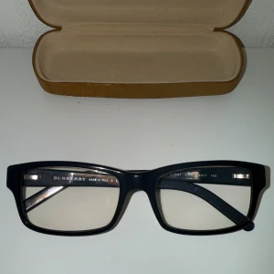 Svarta Burberry glasögon i plast - Snygga svarta glasögon från Burberry med rektangulära bågar i plast. Klassisk och stilren design med diskret logga på insidan av skalmen och på ena utsidan. Tillverkade i Italien och levereras med ett fodral från Timberland.