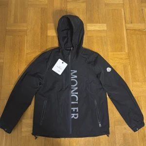 Svart vindjacka från Moncler med huva - Svart vindjacka från Moncler med stor logga framtill, huva och dragkedjor på fickorna. Jackan har Moncler-märke på ärmen och fransk flaggdetalj på ryggen. Tillverkad i polyester och har en clean, sportig look som passar perfekt till streetwear.