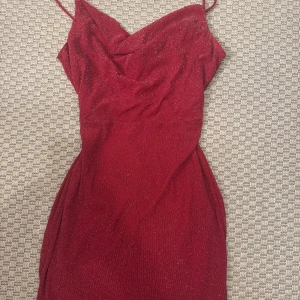 Glittrig vinröd kortklänning - Säljer en snygg vinröd klänning med glittrigt tyg och smala axelband. Klänningen har en draperad halsringning och är figurnära med kort längd, perfekt för att sticka ut. Materialet känns mjukt och har en lyxig finish.