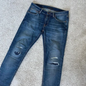 Nudie jeans  - Snygga blå jeans från Nudie Jeans med slitna detaljer. Modellen är skinny lin. Perfekt för dig som gillar en smal passform och coola slitningar. Storlek 28/32 