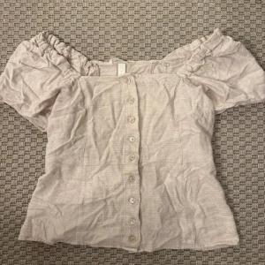 Beige offshoulder blus med knappar - Säljer en söt beige blus i offshoulder-modell med puffiga korta ärmar och knappar hela vägen framtill. Blusen har en lätt rynkad kant upptill och är gjord i ett mjukt bomullsliknande material. Perfekt för dig som gillar romantisk och chill stil.