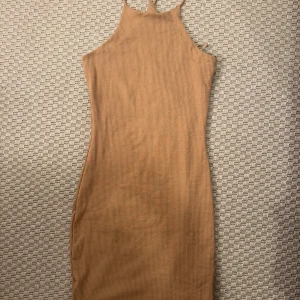 Beige ribbad kortklänning med smala axelband - Säljer en beige ribbad klänning med smala axelband och hög hals. Klänningen är figurnära och har en kort längd som ger en snygg och modern siluett. Perfekt för dig som gillar minimalistisk stil och vill ha något enkelt men ändå trendigt.