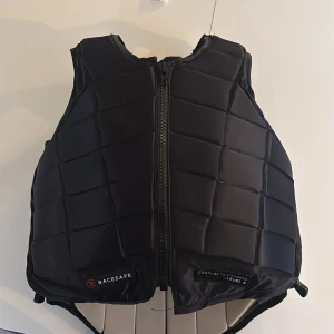 Svart skyddsväst Racesafe RS2010 - Skyddsväst från Racesafe, modell RS2010, i svart med quiltad design och dragkedja framtill. Västen har justerbara sidor med snörning och är tillverkad i syntetmaterial. Perfekt för ridning och ger bra skydd med sin vadderade struktur.