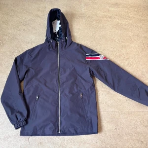 Moncler vindjacka  - Mörkblå vindjacka från Moncler Grenoble med huva, hel dragkedja och två fickor framtill. Ärmen har röd, vit och blå rand samt Moncler-logga. Jackan är tillverkad i polyester och har snygga detaljer som elastiska muddar och innerficka med röd dragkedja.