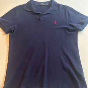 Marinblå pikétröja från Polo Ralph Lauren - Snygg marinblå pikétröja från Polo Ralph Lauren i slim fit-modell. Tröjan har korta ärmar, klassisk krage och knappar framtill. På bröstet finns den  röda Polo-loggan. Storleken är L men passar M perfekt